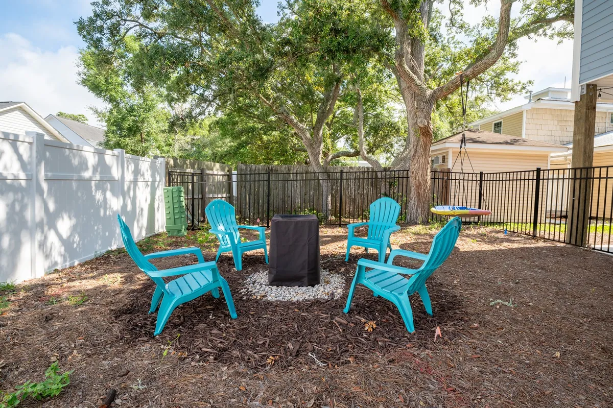 A cozy exterior scene featuring a fire pit encircled by blue chairs, green trees, and a wooden fence. The setting invites small gatherings for fireside chats, evening relaxation, and outdoor games.