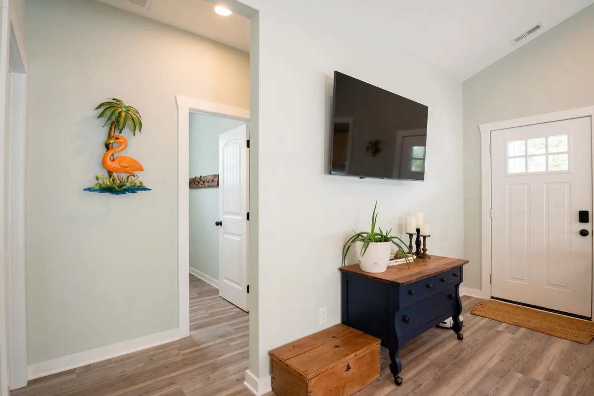 This cheerful entryway scene features flamingo wall art, a practical wooden storage chest, a TV, and a console table with lush plants. The space feels inviting and bright, offering a glimpse of the outdoors through the front door and signaling a welcoming transition from the exterior to the home's interior.