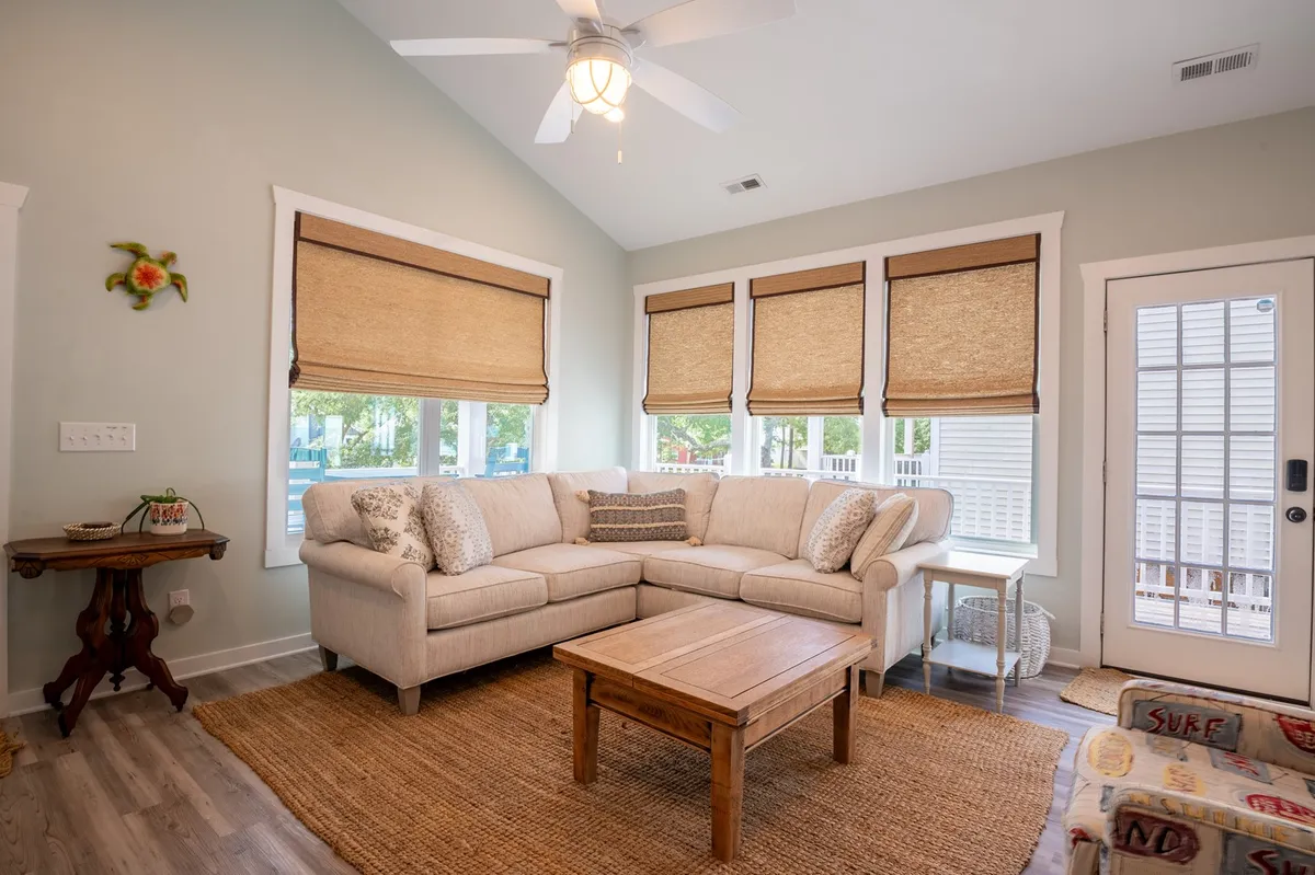 This image displays a cozy living room with a sectional sofa and coffee table positioned by sunlit windows with inviting treatments. The setting is clearly designed for relaxation, reading, or light socializing, with natural light creating a welcoming atmosphere.