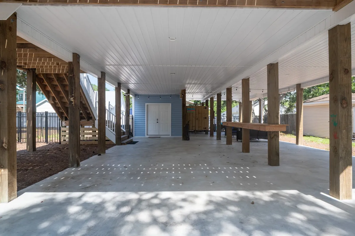An inviting open space beneath the elevated house, featuring sturdy wooden support beams, a long wooden table ideal for group dining, and a visible blue door. The area is shaded, with plenty of room for gatherings, cooking, and for children to play safely. The atmosphere feels casual, welcoming, and perfect for memorable outdoor celebrations and family time.