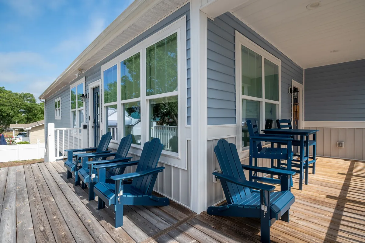 A tranquil exterior deck scene featuring blue Adirondack chairs arranged invitingly, with large windows reflecting the natural light. The wooden deck space is ideal for relaxing, socializing, and enjoying the outdoors. The open setup allows for sunset views and creates a seamless connection to the coastal environment.