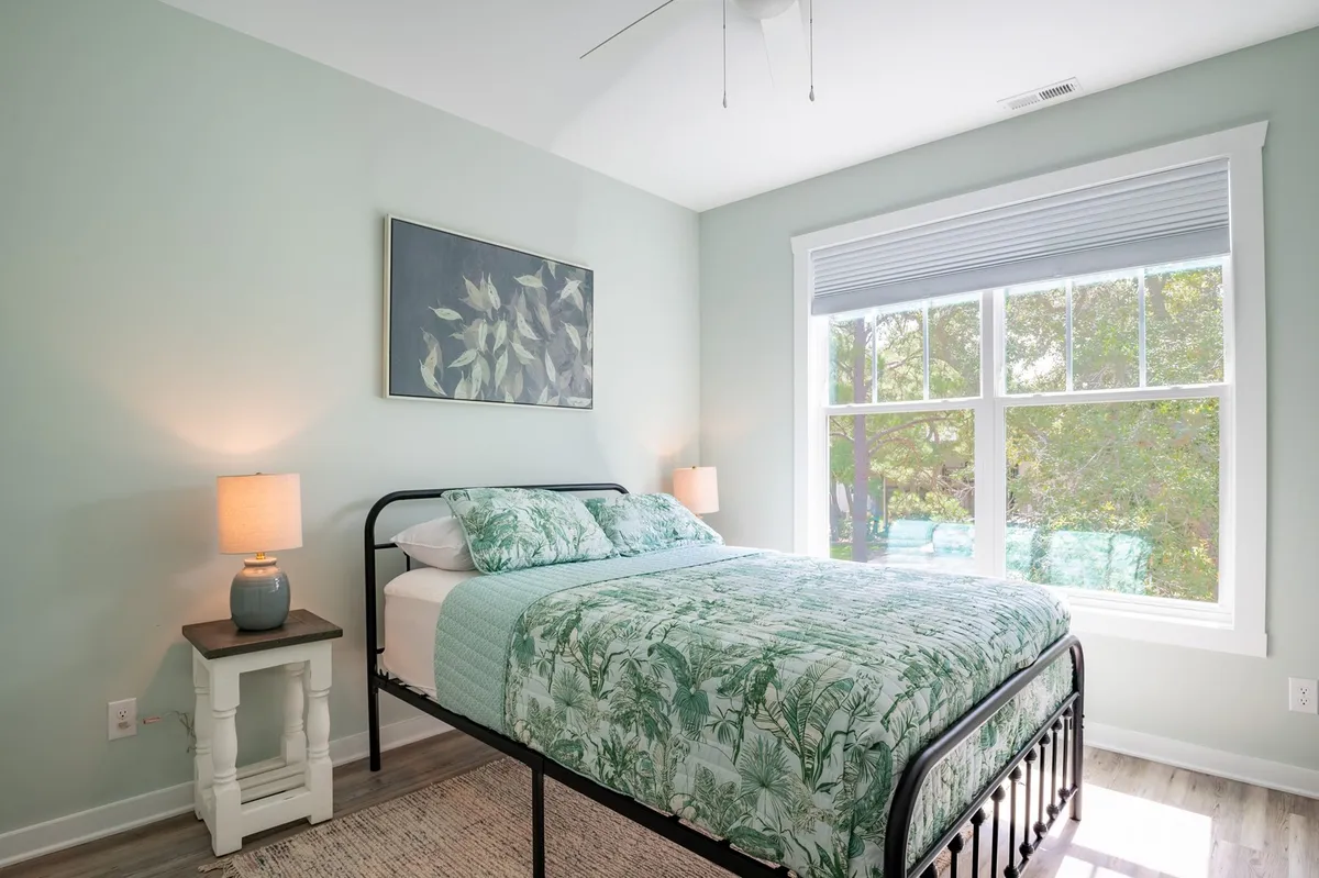 The scene shows a tranquil bedroom with a bed dressed in green bedding, artwork on the wall, and a window letting in natural light. The overall mood is peaceful and restful, making it suitable for both relaxation and quiet activities like reading.