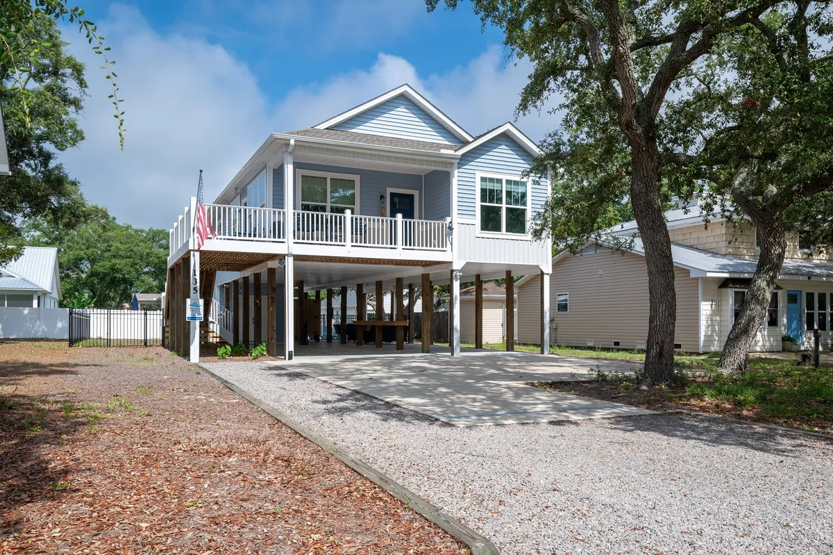 The image features a charming, recently built beach house with a spacious porch draped in gentle shade from mature trees. An American flag adds a festive, welcoming touch, while open space beneath the house and the lush, green lawn suggest ample room for outdoor gatherings, play, and relaxation. The peaceful neighborhood setting is evident, and the porch looks ideal for watching sunsets or hosting family barbecues.