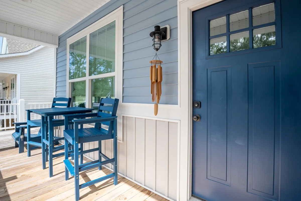 The exterior features a charming blue door, high-top chairs, and wind chimes that sway in the breeze. The setting hints at enjoying morning coffee, greeting neighbors, or soaking in the tranquil coastal atmosphere, all from a vantage point that overlooks the surrounding neighborhood.