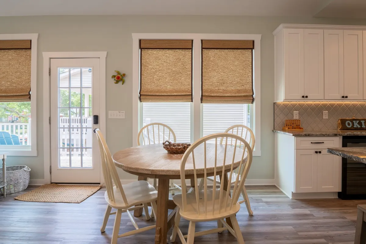 A warm and inviting dining area featuring a round wooden table surrounded by white chairs, kitchen cabinetry in the background, and window treatments that let in natural light. The layout suggests a perfect spot for family meals, morning coffee, or celebrating together.