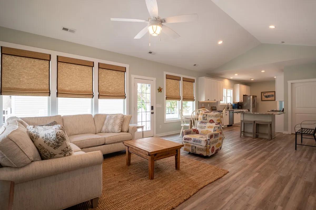 This image features a sunlit open-concept living area. A cozy sofa and decorative armchair frame a coffee table, while the kitchen can be seen in the background, suggesting a seamless flow between spaces. The room is casual and welcoming, with ample natural light streaming in from windows, highlighting the inviting seating and airy atmosphere.