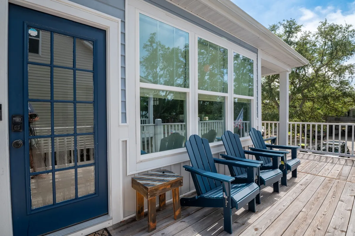 The front porch scene features a blue door, adirondack chairs, and a wooden table, framed by large windows. The setup invites relaxation and socializing, offering a friendly street-facing vantage point that welcomes both guests and pets.