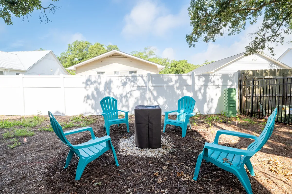 A relaxed outdoor seating area featuring turquoise chairs arranged around a fire pit, all enclosed by a white fence. The setup invites guests to gather for stories, s'mores, or stargazing under the night sky, highlighting the sociable and cozy aspects of the backyard.